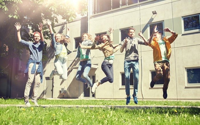 Group of six casually dressed teenagers - three boys and three girls, smiling and jumping in the air outside a building.