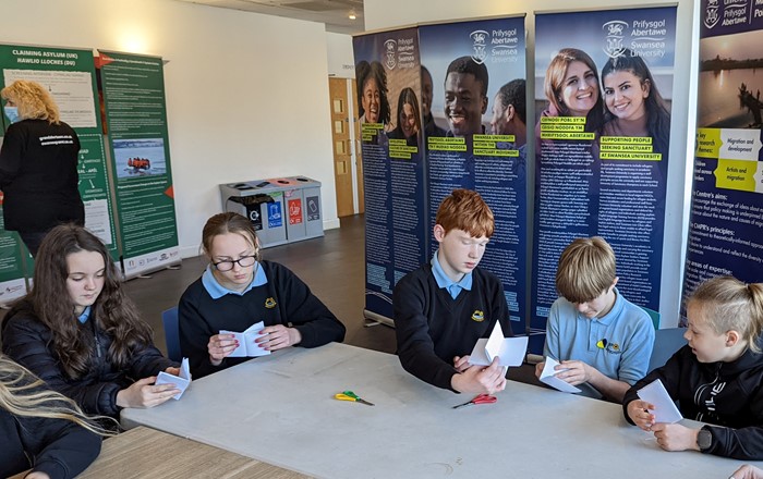 Five school pupils in uniform sitting at a table making things with paper and scissors. Behind them are information panels. Pupils from Ysgol Gyfun Gŵyr in Gowerton taking part in activities at one of the University’s workshops at the exhibition. 