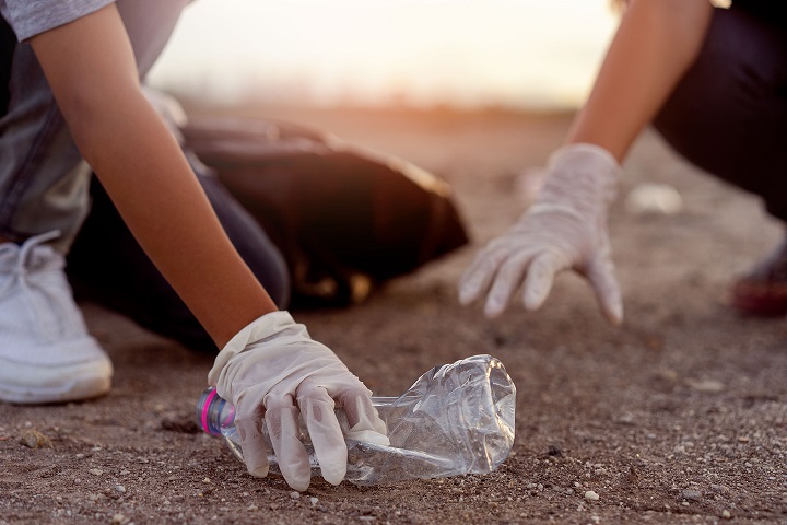two pairs of hands in white gloves on the ground of a beach picking up plastic bottles 