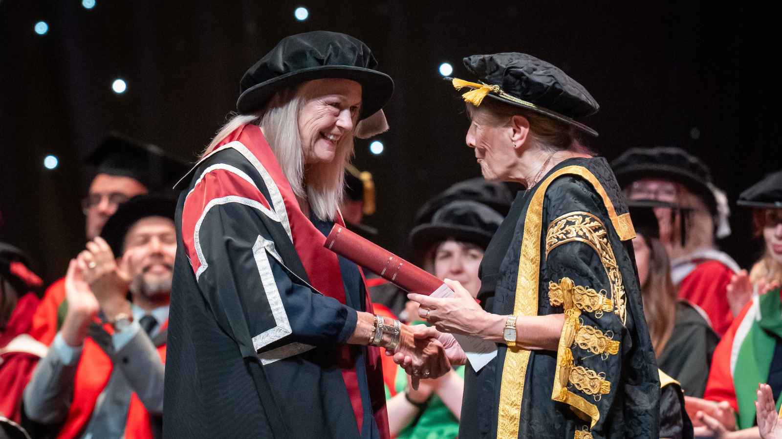 Lady wearing cap and gown, holding a scroll shaking hands with a lady in a Chancellor's Robe