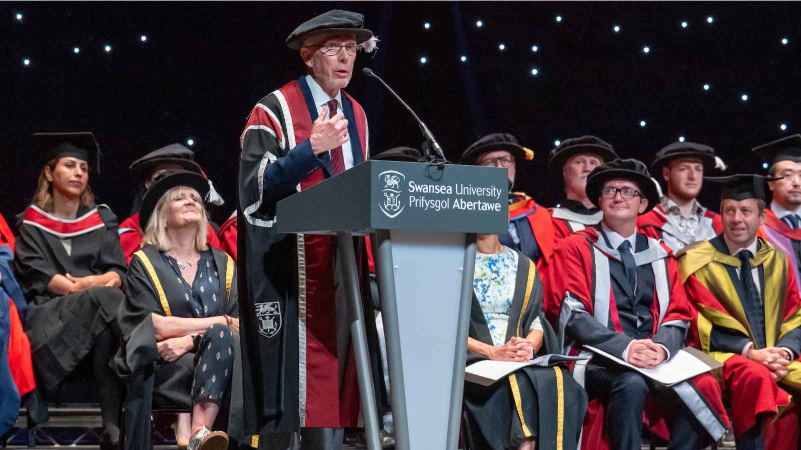 Man wearing cap & gown speaking on stage
