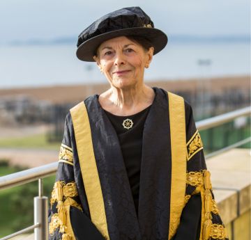 Lady in cap, gown and robes smiling at the camera with the sea behind her.