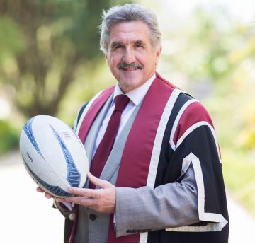 Man in graduation gown holding a rugby ball smiling at the camera