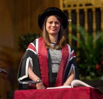 Lady wearing cap & gown standing on a stage behind a table with a book on
