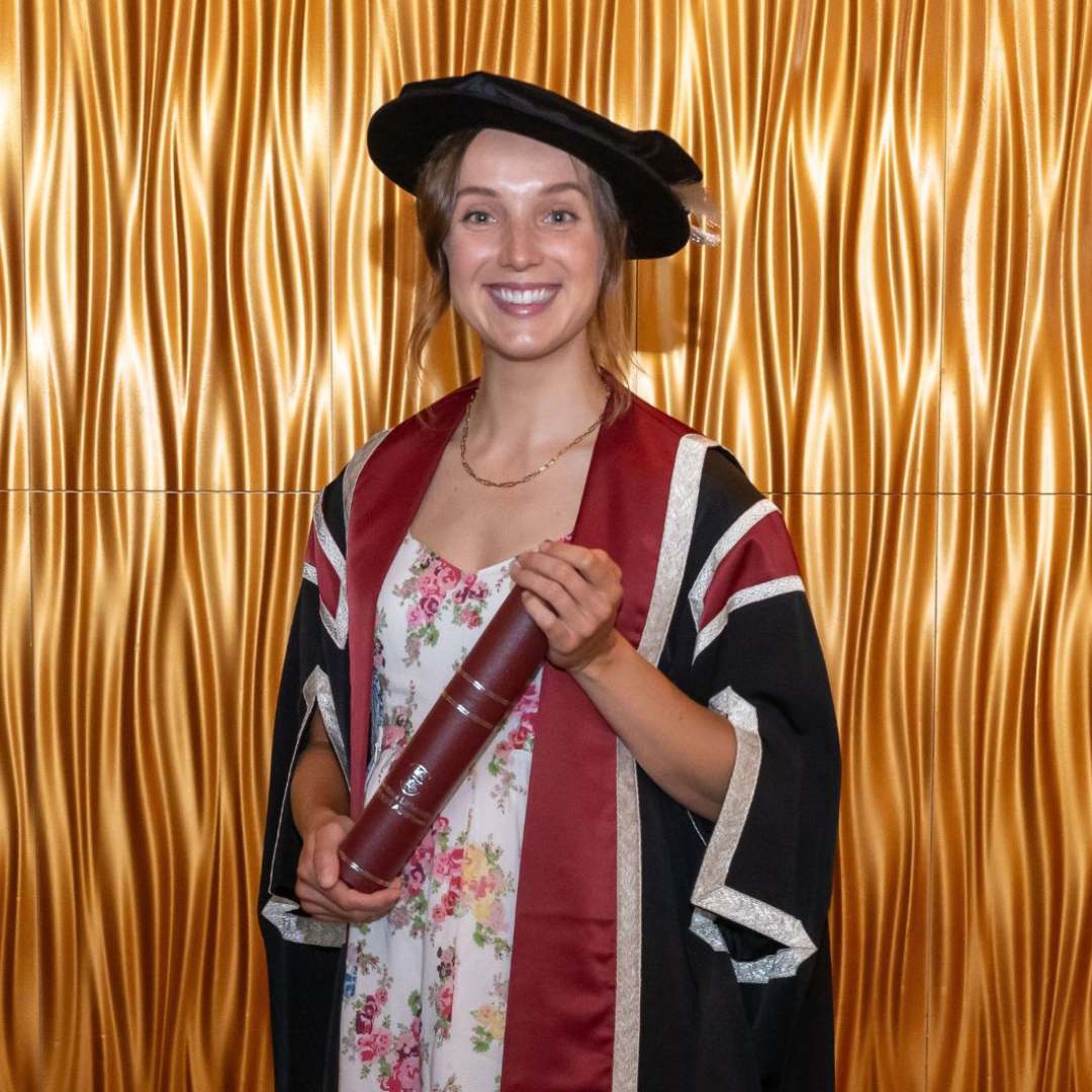 Lady wearing a cap and gown holding a scroll smiling at the camera