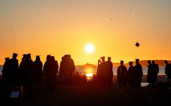 Students standing on a beach during sunset in graduation gowns
