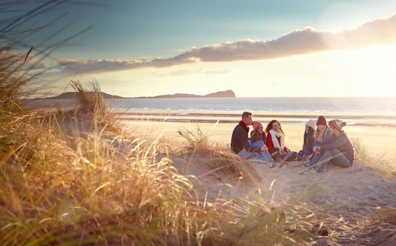 Three students sitting on a Swansea beach smiling & chatting