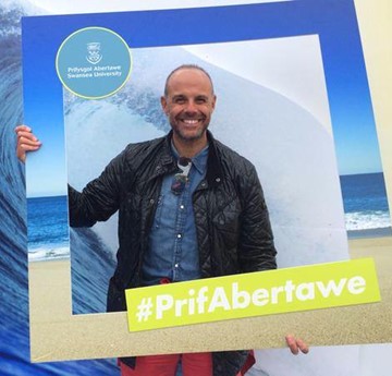 Image of Jason Mohammad stood behind a large frame saying #PrifAbertawe