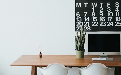 An image of a desk with a computer, chair and a calendar on the wall. 