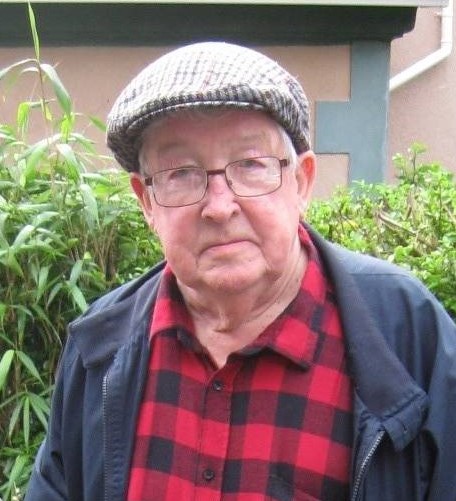 Alun Rees in a red check shirt and flat cap in front of a green leafy hedge and a house