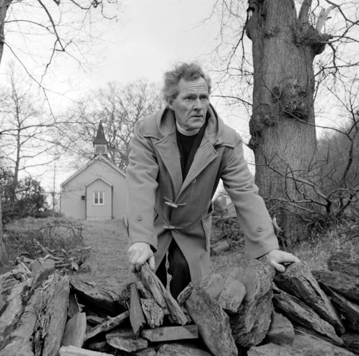 R S Thomas in a duffel coat leaning on a stone wall with a church or chapel in the background