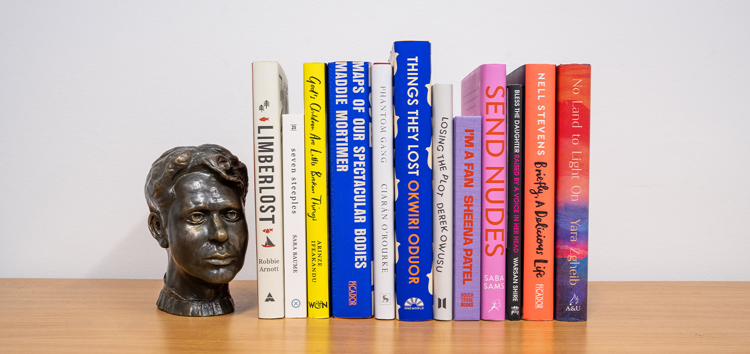 Image of spines of the 12 longlisted books on bookshelf alongside bronze bust of Dylan Thomas
