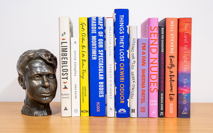 Image of spines of the 12 longlisted books on bookshelf alongside bronze bust of Dylan Thomas