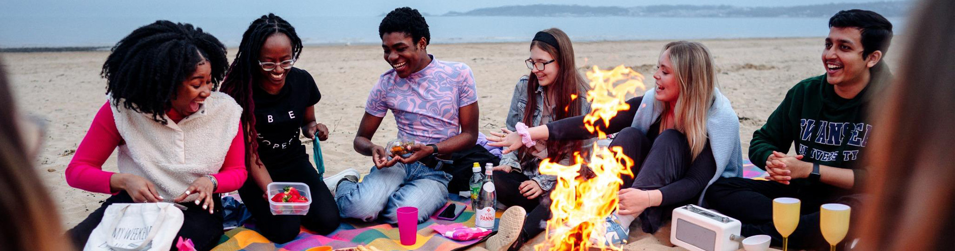 Myfyrwyr yn sgwrsio yn yr haul y tu allan i adeiladau ar gampws y Bae