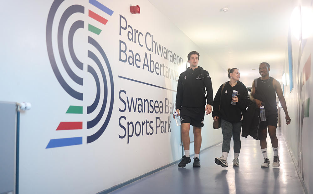 Tri myfyriwr yn gwisgo cit chwaraeon Prifysgol Abertawe yn cerdded ar hyd y coridor i’r Neuadd Chwaraeon a’r gampfa ar Lôn Sgeti – mae logo mawr Parc Chwaraeon Bae Abertawe ar wal y coridor
