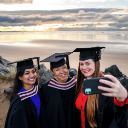 Delwedd o raddedigion yn tynnu hunlun ar y traeth wrth ymyl Campws y Bae.