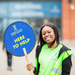 Delwedd o Llysgenhadur Myfyrwyr yn dal arwydd 'Yn Yma i Helpu' ar Gampws y Bae.