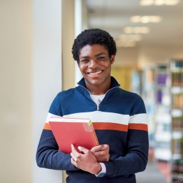 Delwedd o fyfyriwr yn sefyll yn y llyfrgell yn dal llyfrau.