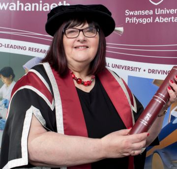 Menyw mewn gwisg graddio yn dal scroll ac yn gwenu wrth y camera