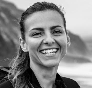 Menyw yn sefyll o flaen y mor yn gwenu i'r camera