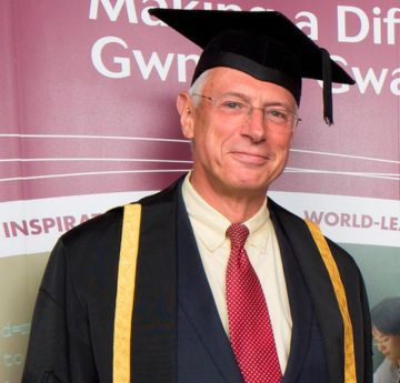 Dyn mewn gwisg graddio yn gwenu i'r camera