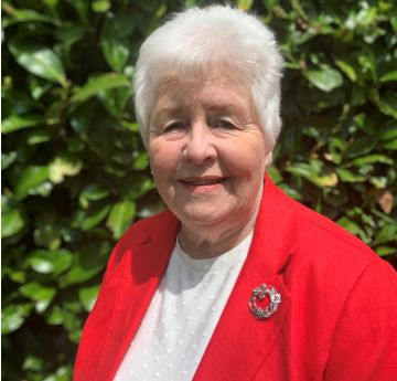 Menyw mewn siaced goch yn sefyll o flaen perth yn gwenu i'r camera