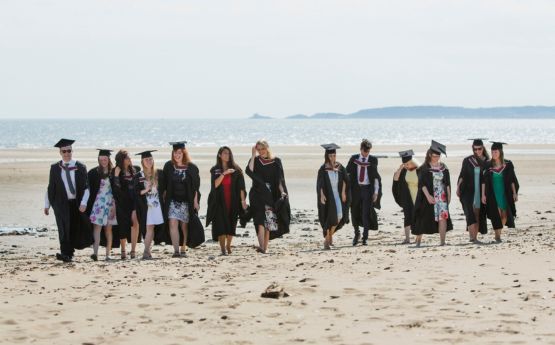 Myfyrwyr yn eu gwisgoedd graddio yn cerdded ar y traeth