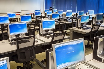 A classroom with desks and PCs. All the PCs are switched on and showing the login screen.