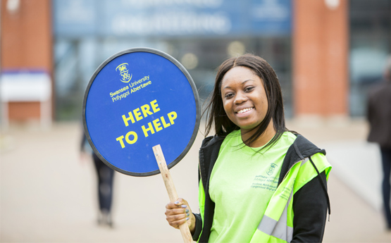 Myfyriwr yn dal lolipop ar diwrnod agored sy'n darllen 'yma i helpu'