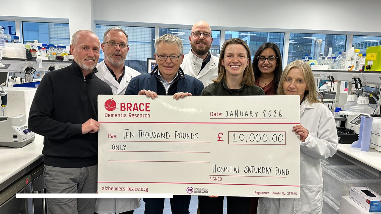 Grŵp o saith o bobl yn sefyll mewn labordy yn dal siec fawr