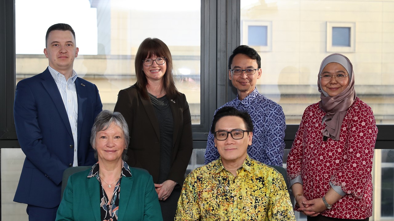 Chwech o bobl wedi'u darlunio dan do, pedwar yn sefyll, dau yn eistedd o'u blaenau wrth fwrdd.