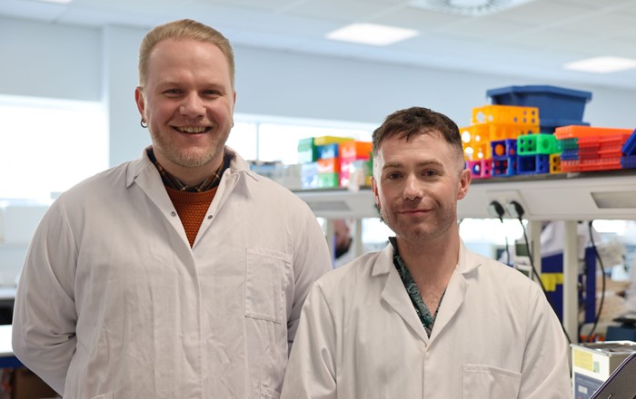 Dau ddyn yn gwisgo cotiau gwyn yn sefyll mewn labordy.