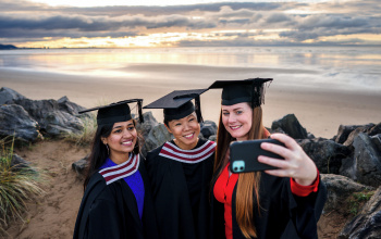 Tair menyw mewn gwisg graddio yn cymryd hunlun ar y traeth