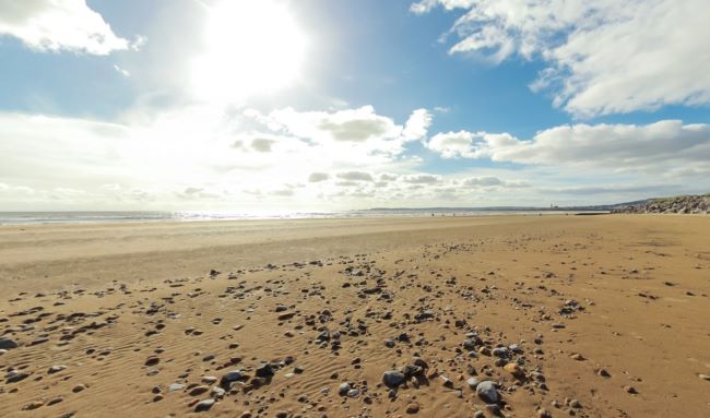 Traeth tywodlyd llydan gyda cherrig mân gwasgaredig o dan awyr lachar chymylog