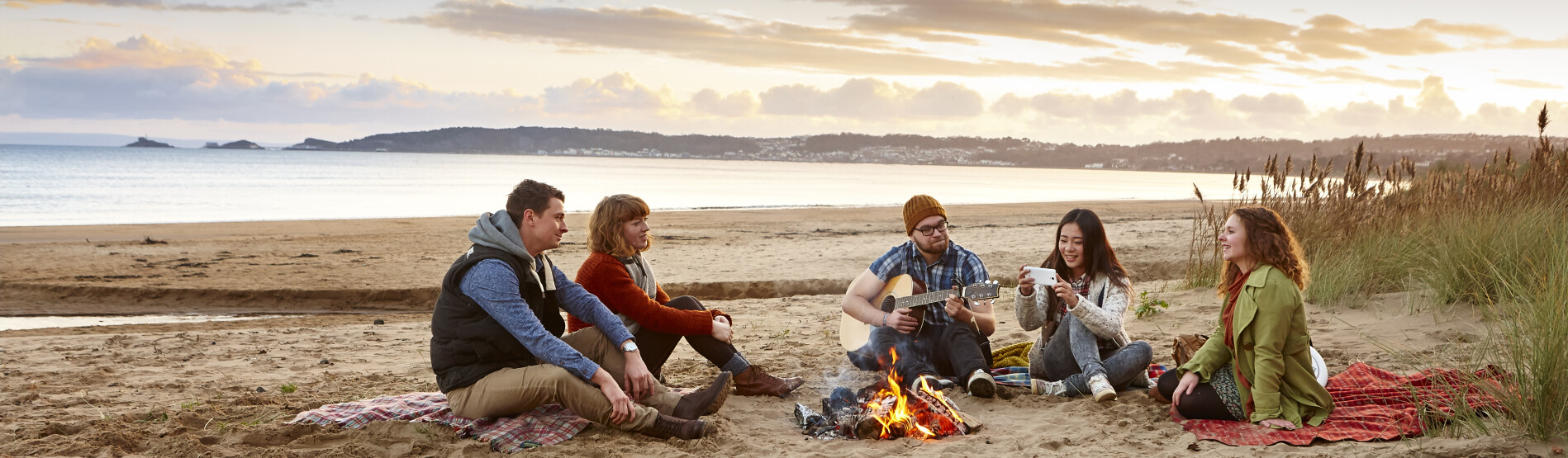 Students playing guitar around a campfire on the beach.
