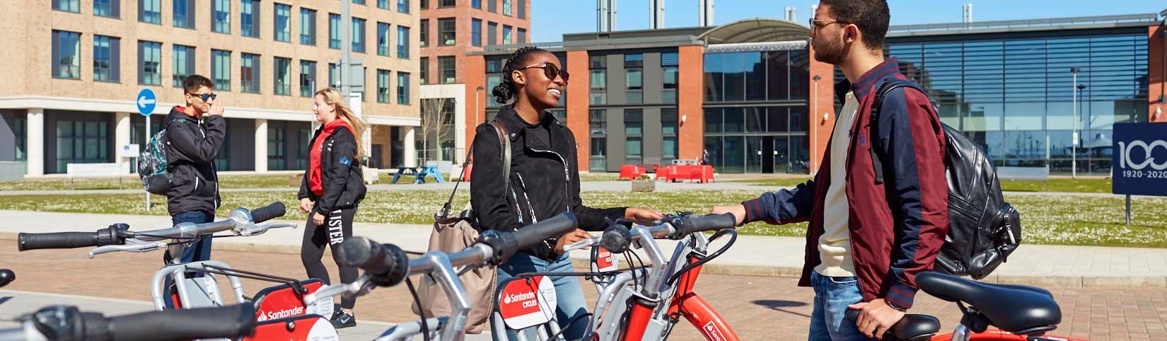 Two students chatting at the Santander Cycles docking station at the Bay Campus with Engineering Central building in the background.