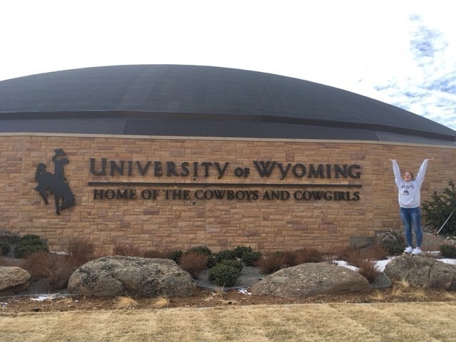 Student standing in front of a wall with 'University of Wyoming: Home of the cowboys and cowgirls' on it with a logo of a cowboy on a horse