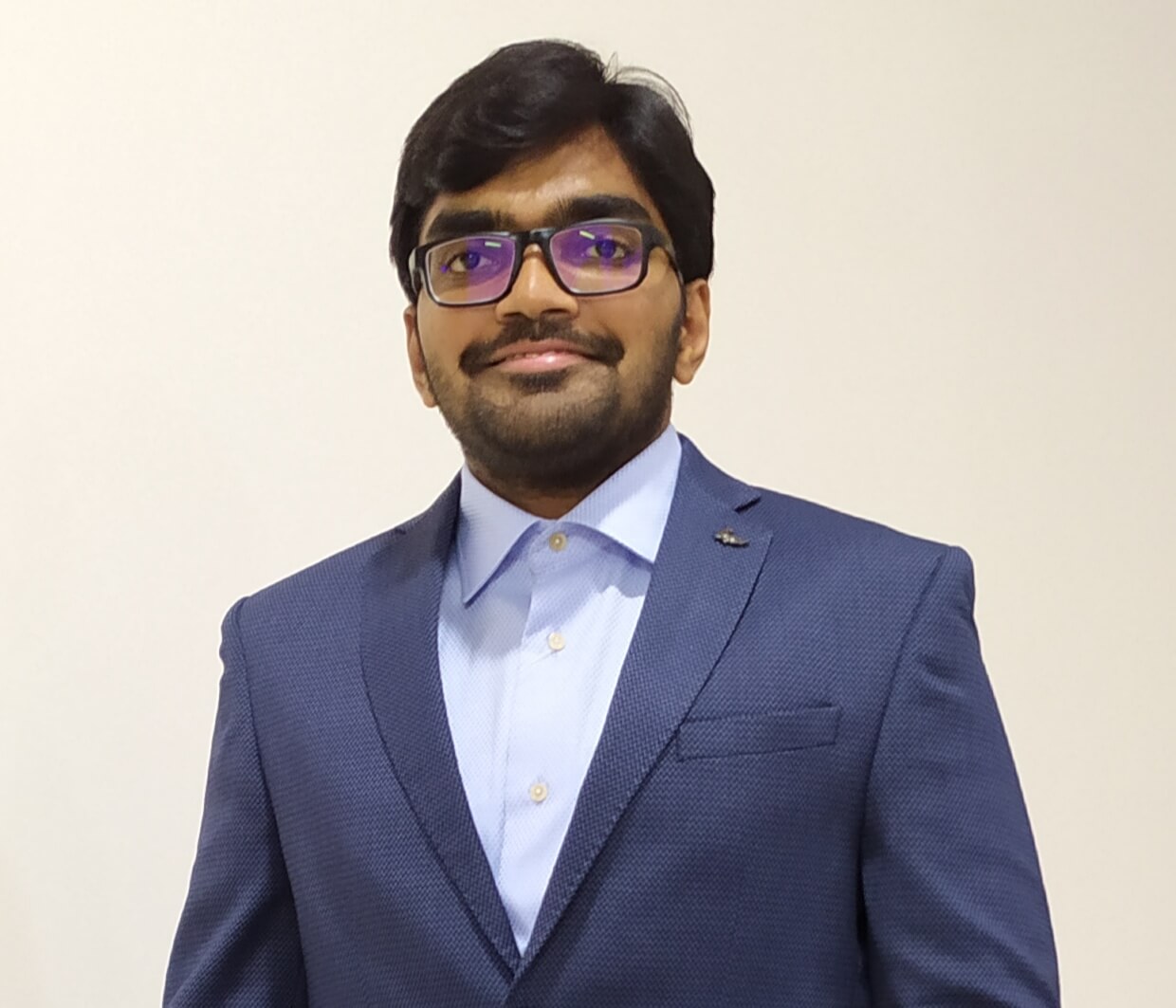 A portrait of Sai, in a smart navy suit smiling at the camera on a cream background.