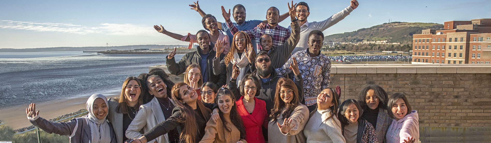 Photo of the 2021 Chevening Scholars waving and posing on a balcony over looking the Bay Campus Beach and buildings 