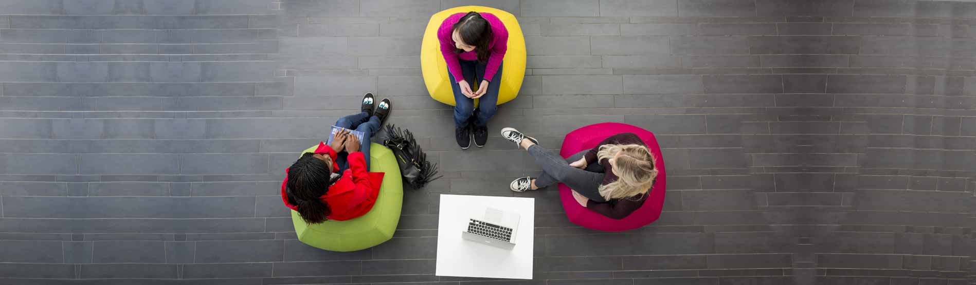 an aerial shot of three students talking, sitting on brightly coloured chairs with a grey slate floor