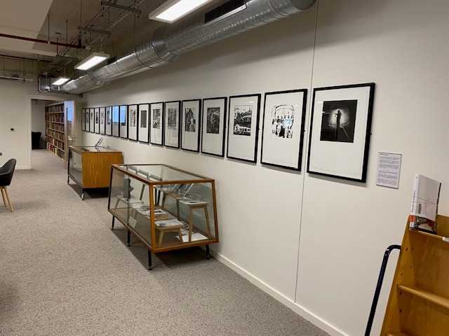 A row of black frames on a white wall containing Black and white artwork from the South Wales Miners' Library Collection
