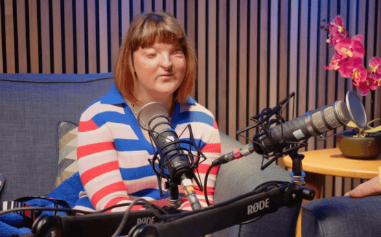 A woman sitting on a grey sofa speaking into a microphone for a podcast.