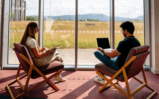 Two students using a laptop and reading a book in chairs in the library next to a wide window