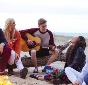 Students on the beach around a campfire with a boy playing a guitar. 