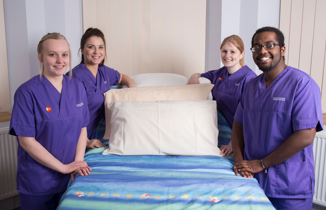 Four nursing students in scrubs surround a hospital bed with pillows