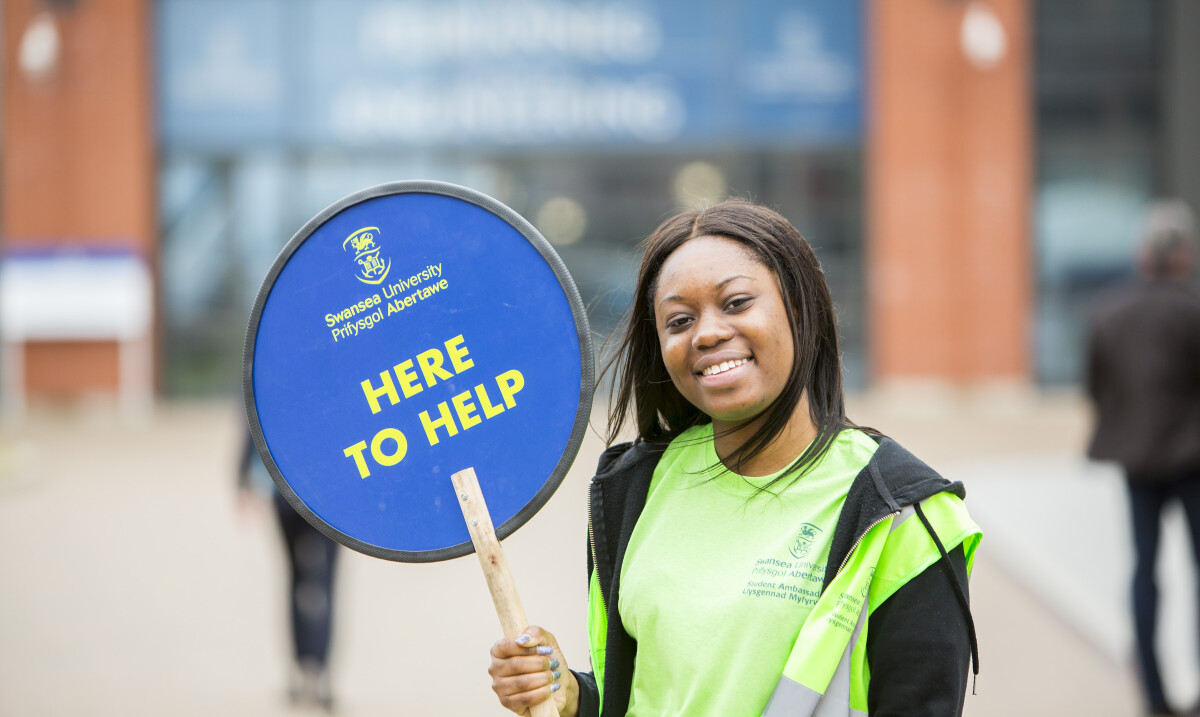 Image of a student ambassador holding up a here to help sign at an open day 