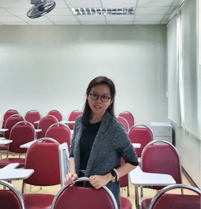 Student wearing glasses, standing behind chair and smiling at camera.