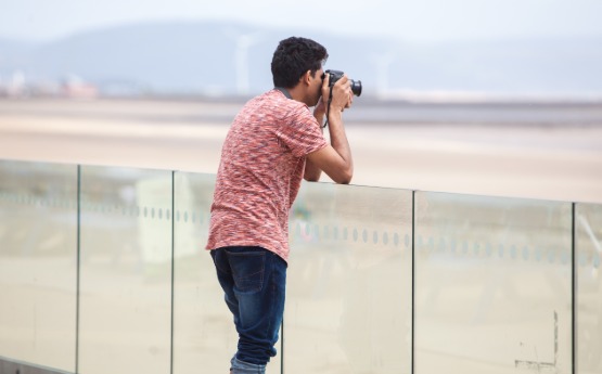 Male student leaning on wall outside 360 and taking a photograph with a camera