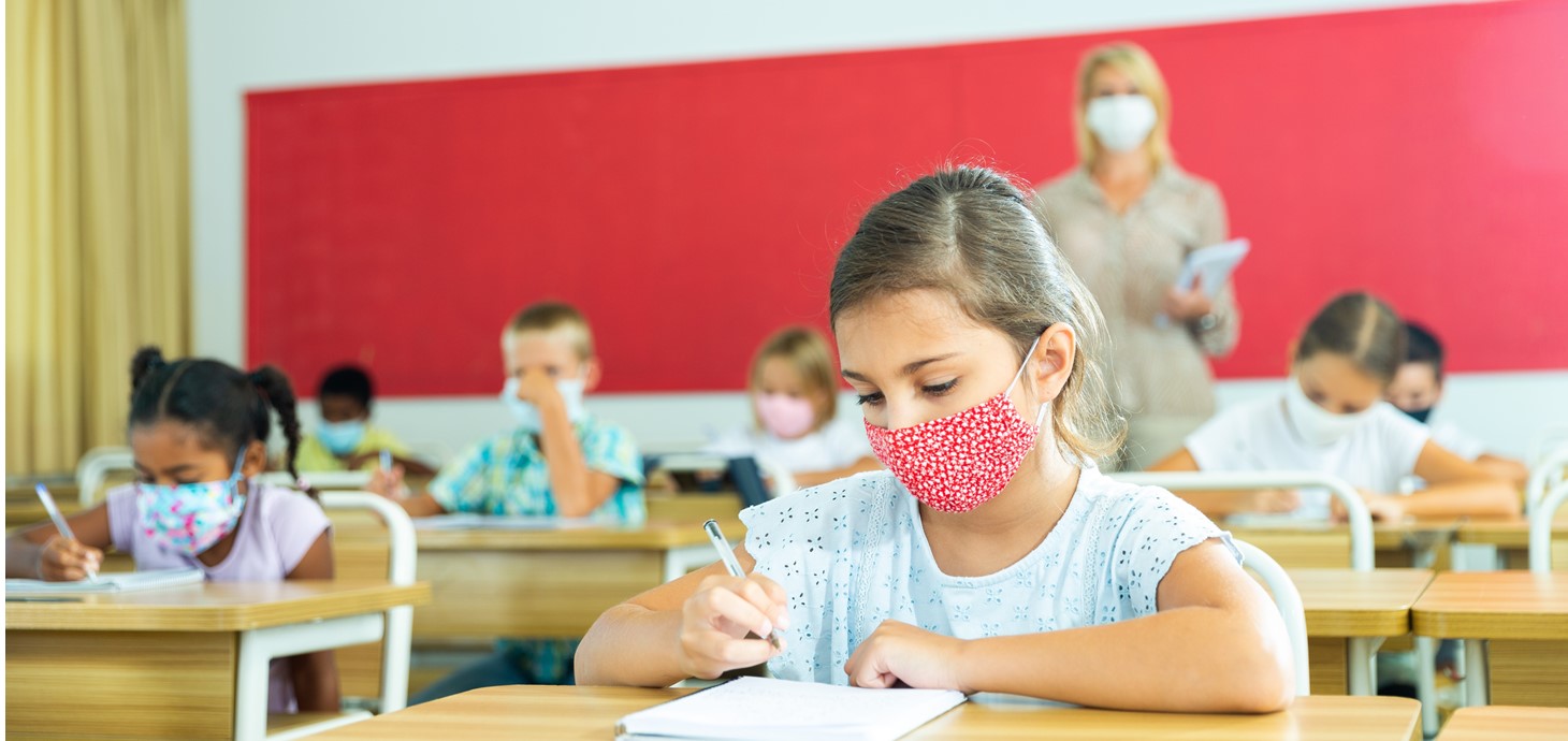 A young schoolgirl wearing a face mask sits writing at her desk, with the teacher and other pupils in the background. 