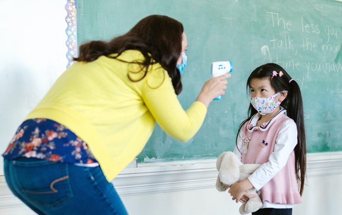 Adult teacher wearing a mask taking temperature of primary school pupils, also wearing face mask, in a classroom.